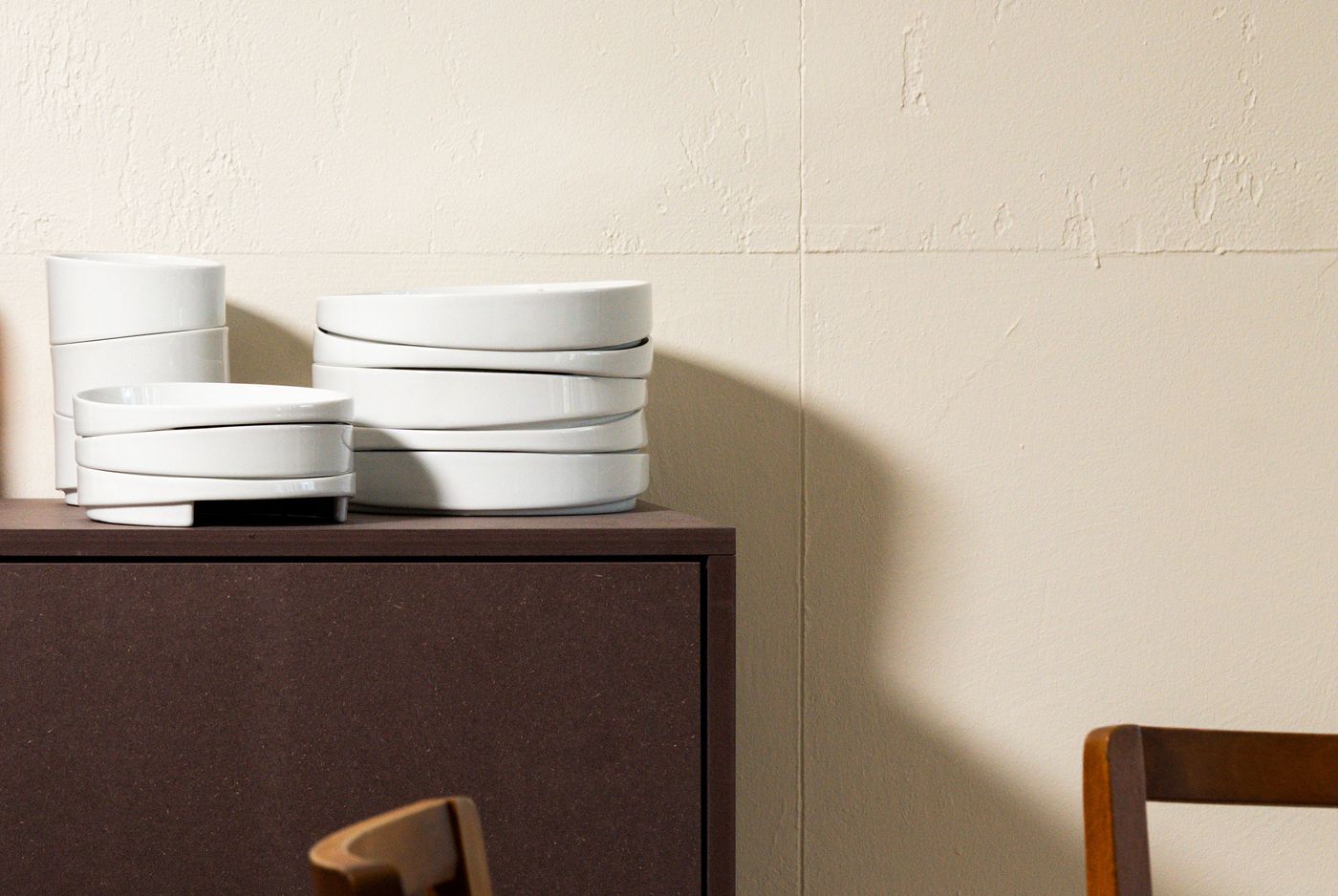 A cozy corner with minimalist stacked white ceramics placed on a dark brown cabinet against a neutral wall.