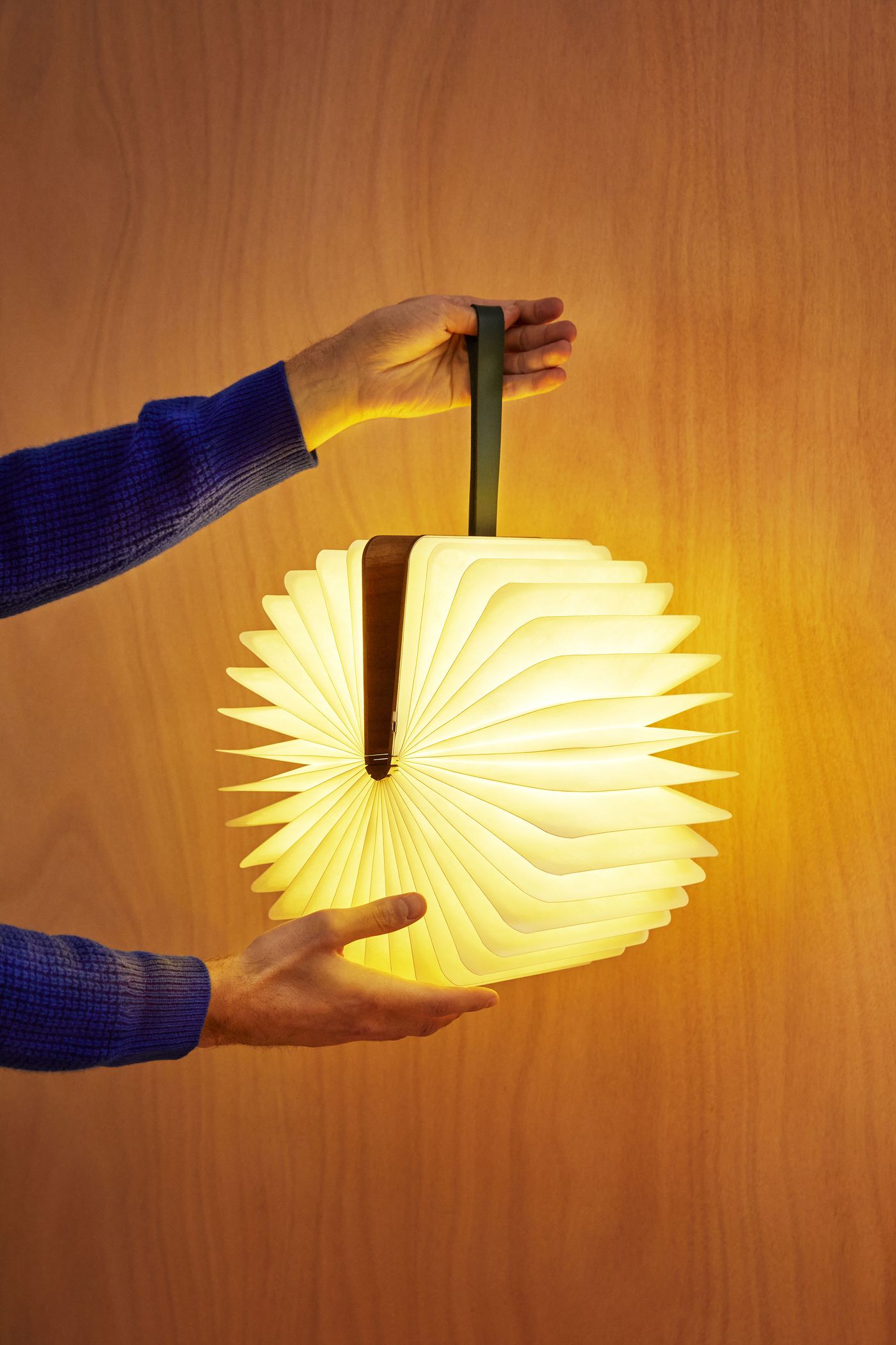 A person holds a fully expanded circular book lamp with a handle, its pleated pages glowing in soft yellow against a wooden wall.
