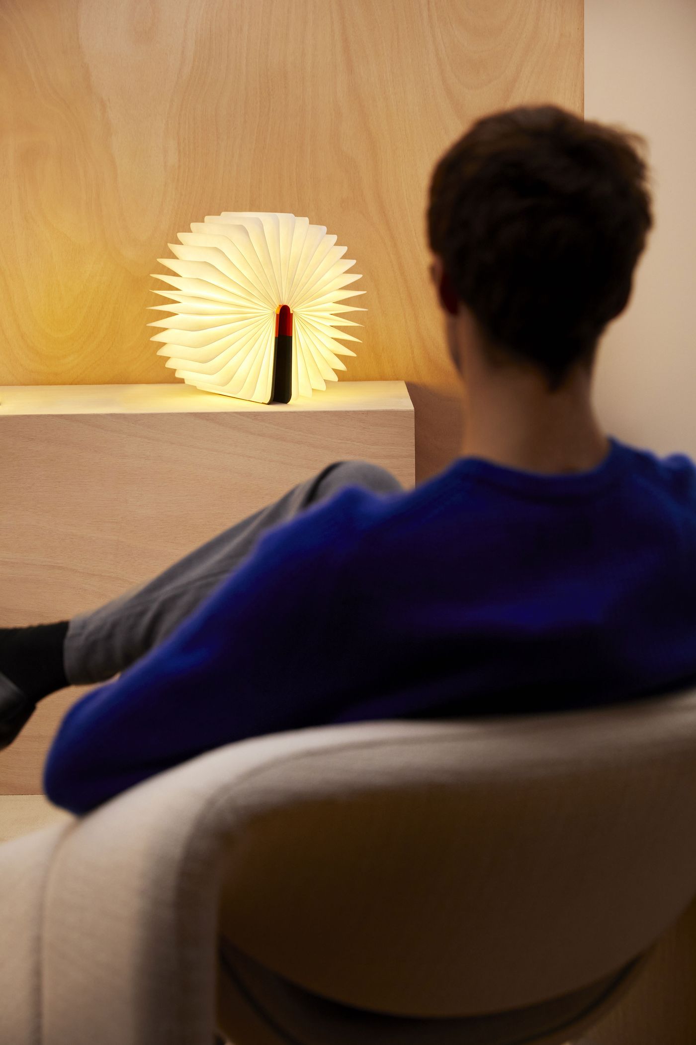 A person seated in an armchair gazes at a luminous sculptural book lamp on a pedestal, shaped like a radiant sunburst. A person seated in an armchair gazes at a luminous sculptural book lamp on a pedestal, shaped like a radiant sunburst.