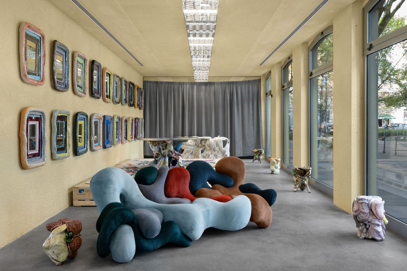 A gallery-like interior filled with playful, surrealist furniture by Misha Kahn, including an undulating, multicolored velvet lounge piece at the center, organic ceramic sculptures scattered along the floor, and a wall lined with irregularly framed mirrors in vibrant hues—eccentric, tactile, and joyfully maximalist. A gallery-like interior filled with playful, surrealist furniture by Misha Kahn, including an undulating, multicolored velvet lounge piece at the center, organic ceramic sculptures scattered along the floor, and a wall lined with irregularly framed mirrors in vibrant hues—eccentric, tactile, and joyfully maximalist.