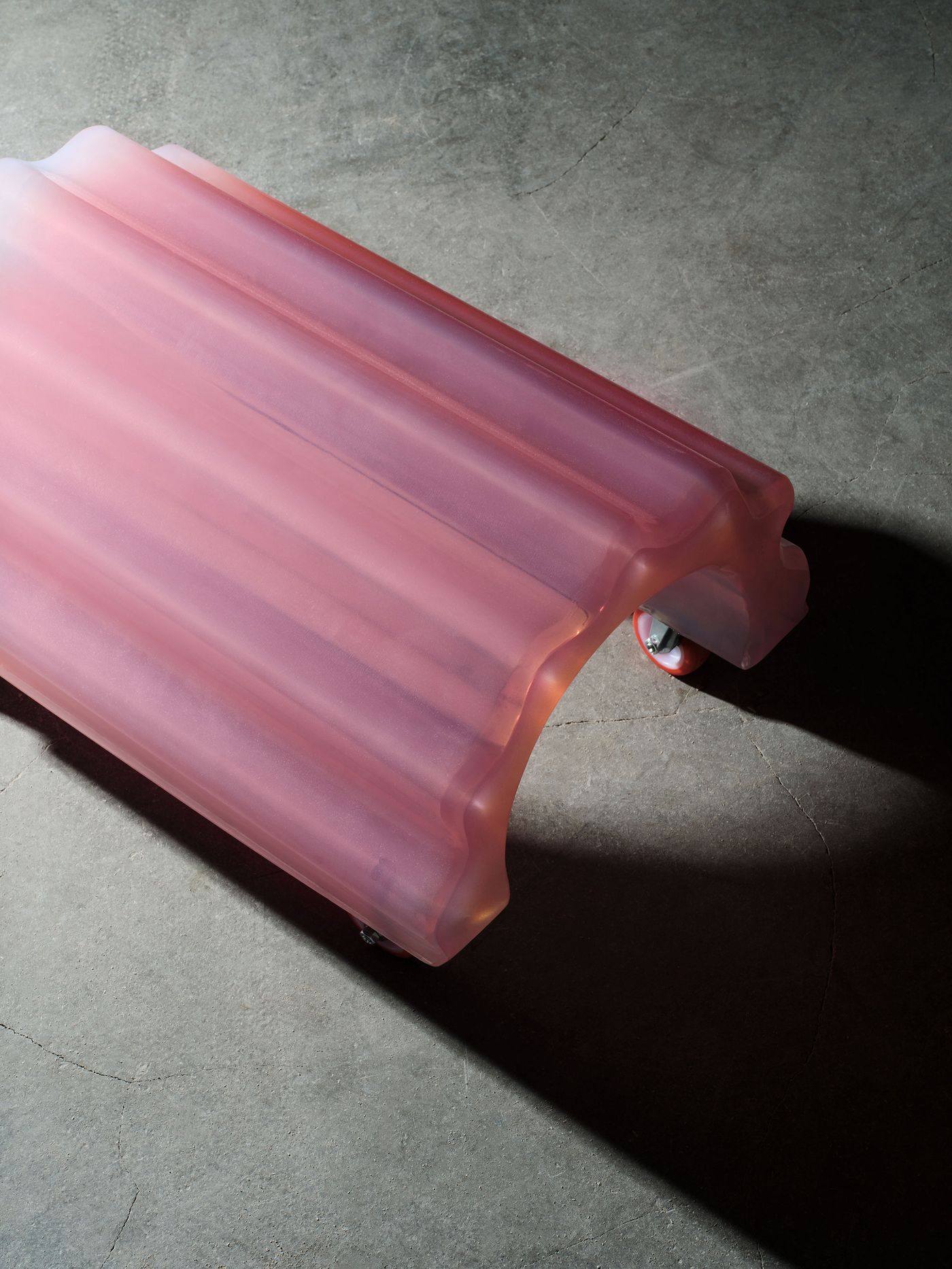A translucent pink stool with a fluted, arched form and built-in wheels, made from resin-like material, its jelly-like surface catching soft light on a concrete floor—playful, futuristic, and oddly tactile. A translucent pink stool with a fluted, arched form and built-in wheels, made from resin-like material, its jelly-like surface catching soft light on a concrete floor—playful, futuristic, and oddly tactile.