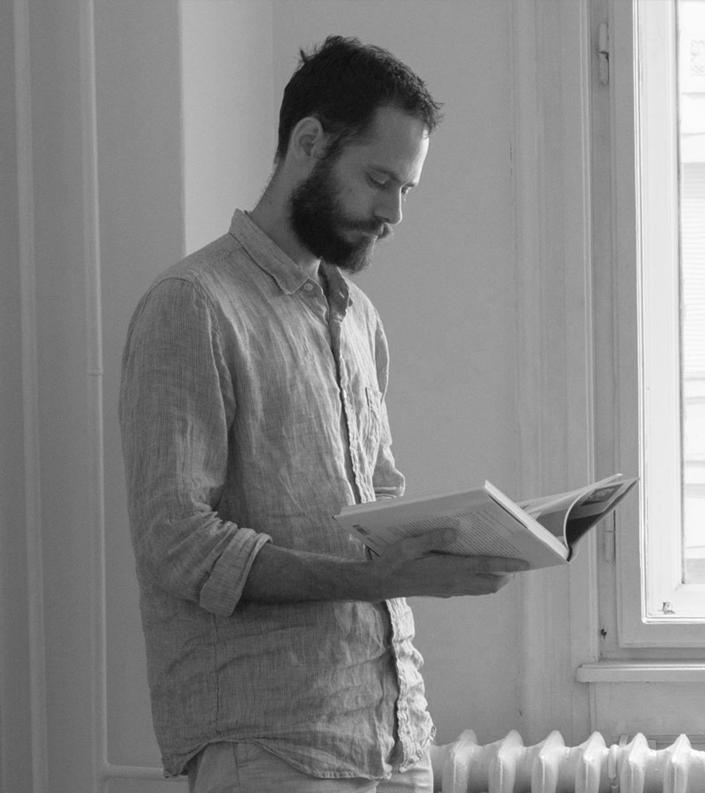 A black and white portrait captures collector Gonçalo Campos, a bearded man, standing by a window and intently reading a book. His relaxed posture and simple linen shirt evoke a contemplative, authentic mood, reflecting Yatzer's appreciation for individuals deeply connected to the art of living beautifully.