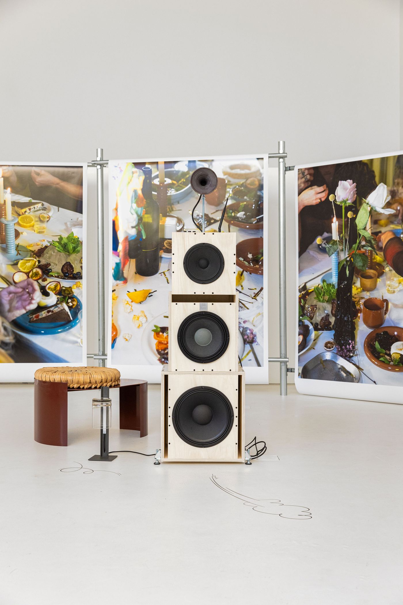A striking display from the "Sobre Mesa" exhibition by Aires Mateus features a tall, stacked audio system with large speakers, positioned on a light floor with subtle line drawings. Behind it, three large photographic prints depict vibrant, richly laden dining tables. The composition creates an intriguing juxtaposition between functional design, artistic photography, and a playful, immersive atmosphere.