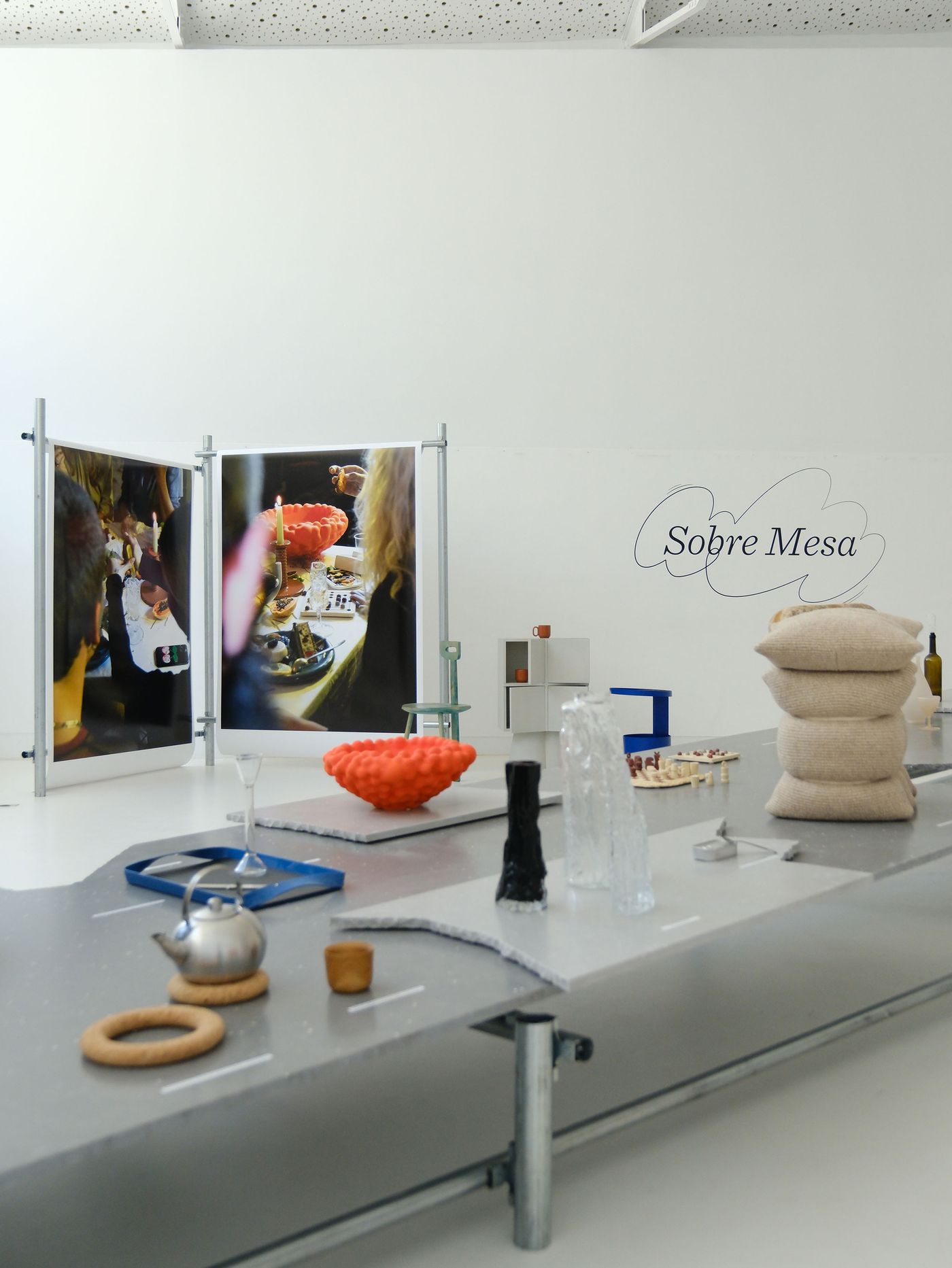 An art installation featuring a gray, undulating surface reminiscent of a road, displaying various sculptural objects, including an orange textured piece and a large stack of beige cushions. In the background, two large panels display vibrant photographic prints of a lively table setting. Titled "Sobre Mesa" by Aires Mateus, the scene creatively explores domesticity and artistry through unconventional design elements.