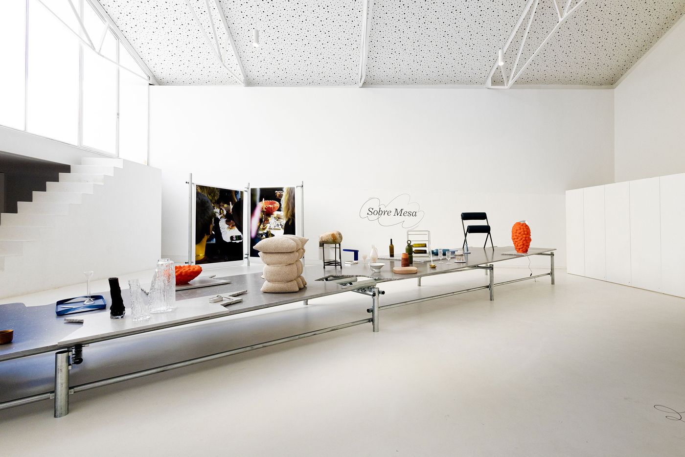 The "Sobre Mesa" exhibition by Arquivo Aires Mateus features a long, low display table showcasing a diverse collection of contemporary tableware and ceramic objects in muted and vibrant tones. Behind it, large photographic panels depict lively dining scenes, creating a dynamic interplay between the exhibited objects and their potential use, set within a spacious, modern gallery.