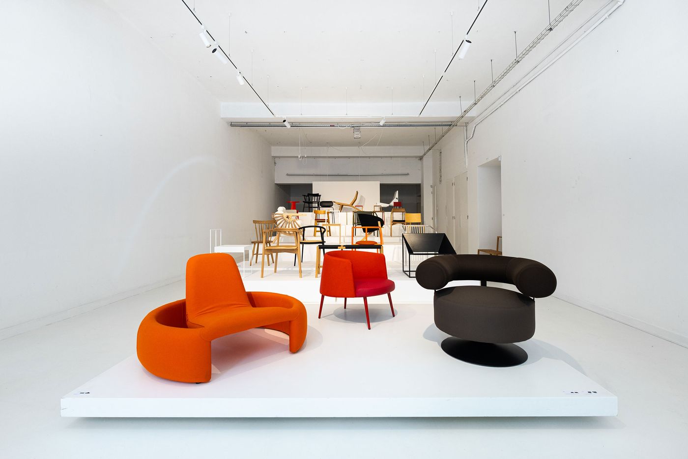 A pristine white exhibition hall showcases an eclectic array of modern chairs, with a vibrant orange modular seat, a red accent chair, and a dark brown rounded armchair prominent in the foreground. In the background, a diverse collection of wooden and contemporary chairs continues the display. The clean, open space with track lighting emphasizes each piece's unique form, typical of Branca Lisboa's curated selection.