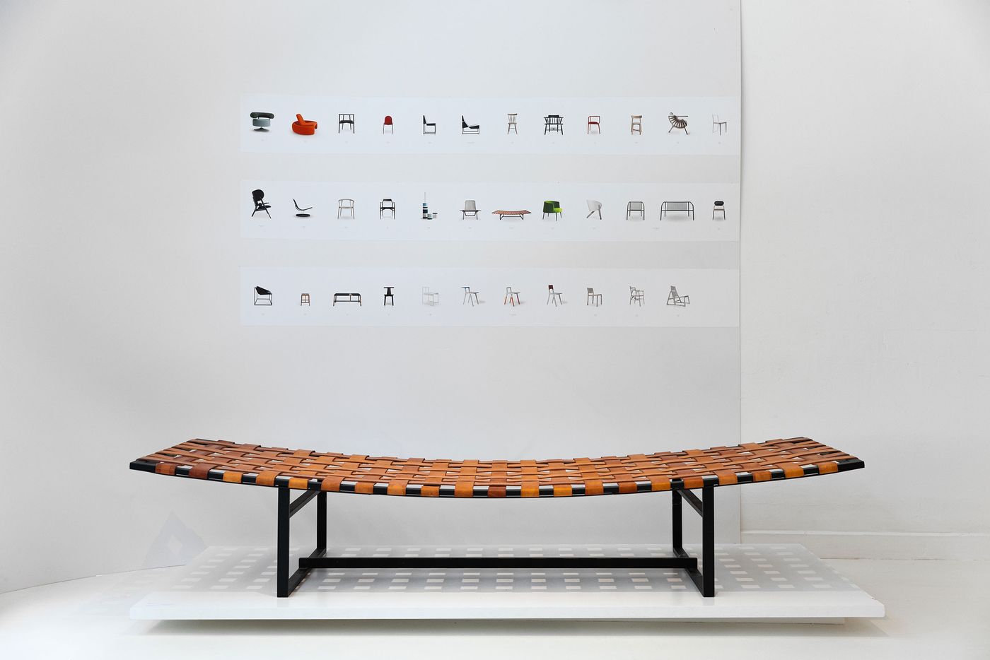 A contemporary bench with a curved, woven tan leather seat and a minimalist black metal frame sits centrally in a white exhibition space. Behind it, a wall display features three rows of diverse chair silhouettes, creating a visual narrative of form and function. The composition highlights classic design principles and craftsmanship, typical of Branca Lisboa's focus on timeless pieces.