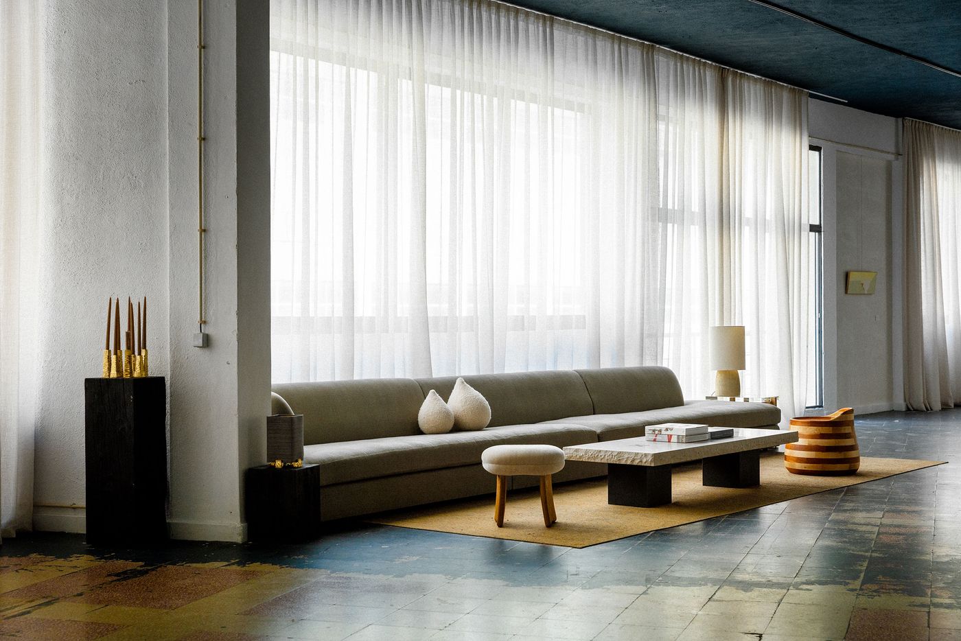 A minimalist Hamrei living space features a long, low-slung grey sofa facing a rough-hewn stone coffee table on a neutral rug. Floor-to-ceiling sheer white curtains create a soft, diffused light, contrasting with the dark industrial ceiling and concrete floor. Gold accents, a sculptural lamp, and a striped stool add subtle warmth, creating a serene, contemporary ambiance.