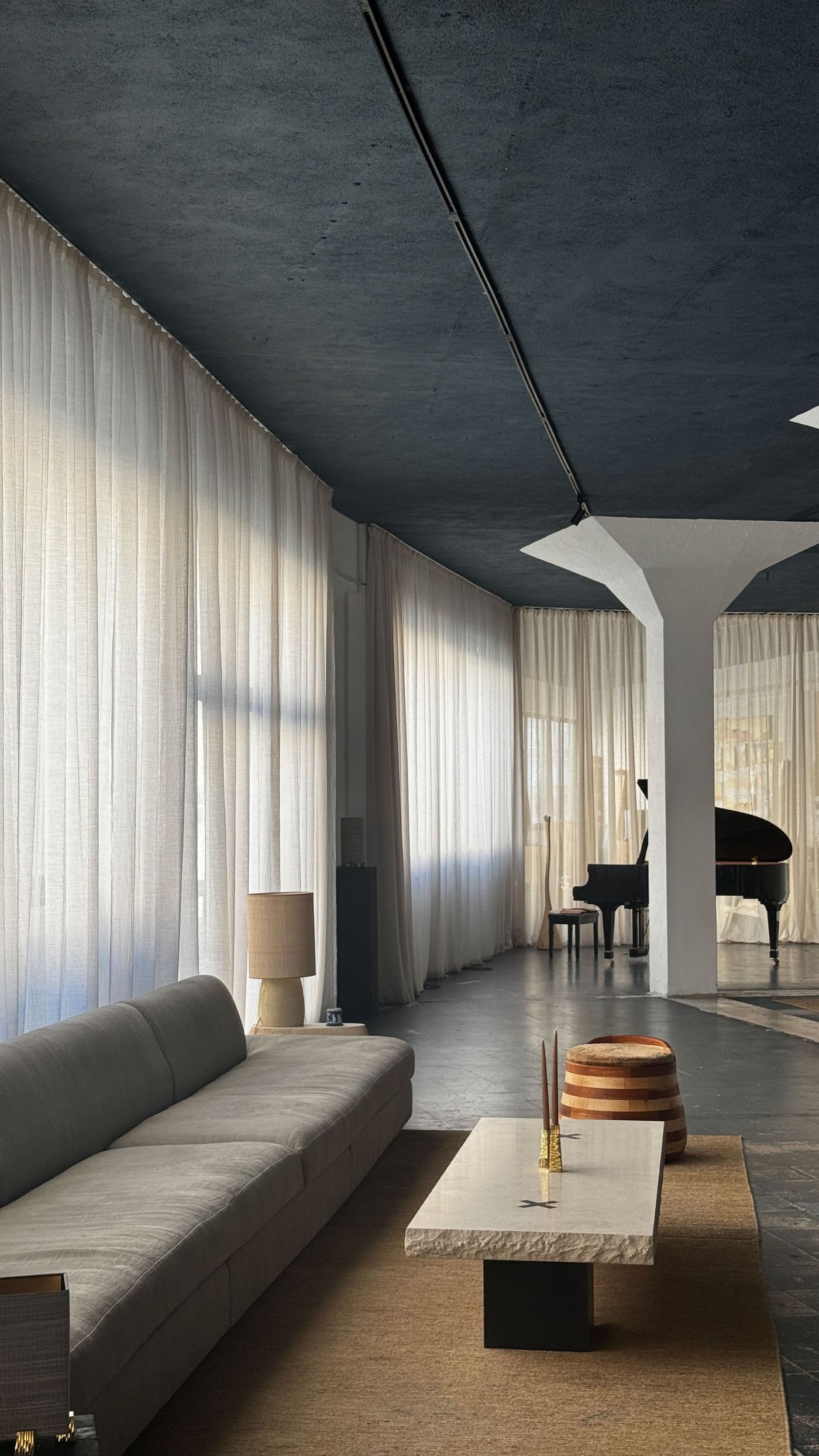 A grand piano subtly emerges from the background of a refined Hamrei living space, characterized by a deep grey ceiling, fluid white sheer curtains, and a long, low-slung beige sofa. The concrete floor and minimalist furnishings create a contemplative atmosphere, where the interplay of light and shadow, typical of a Hamrei design, evokes a sense of understated elegance.