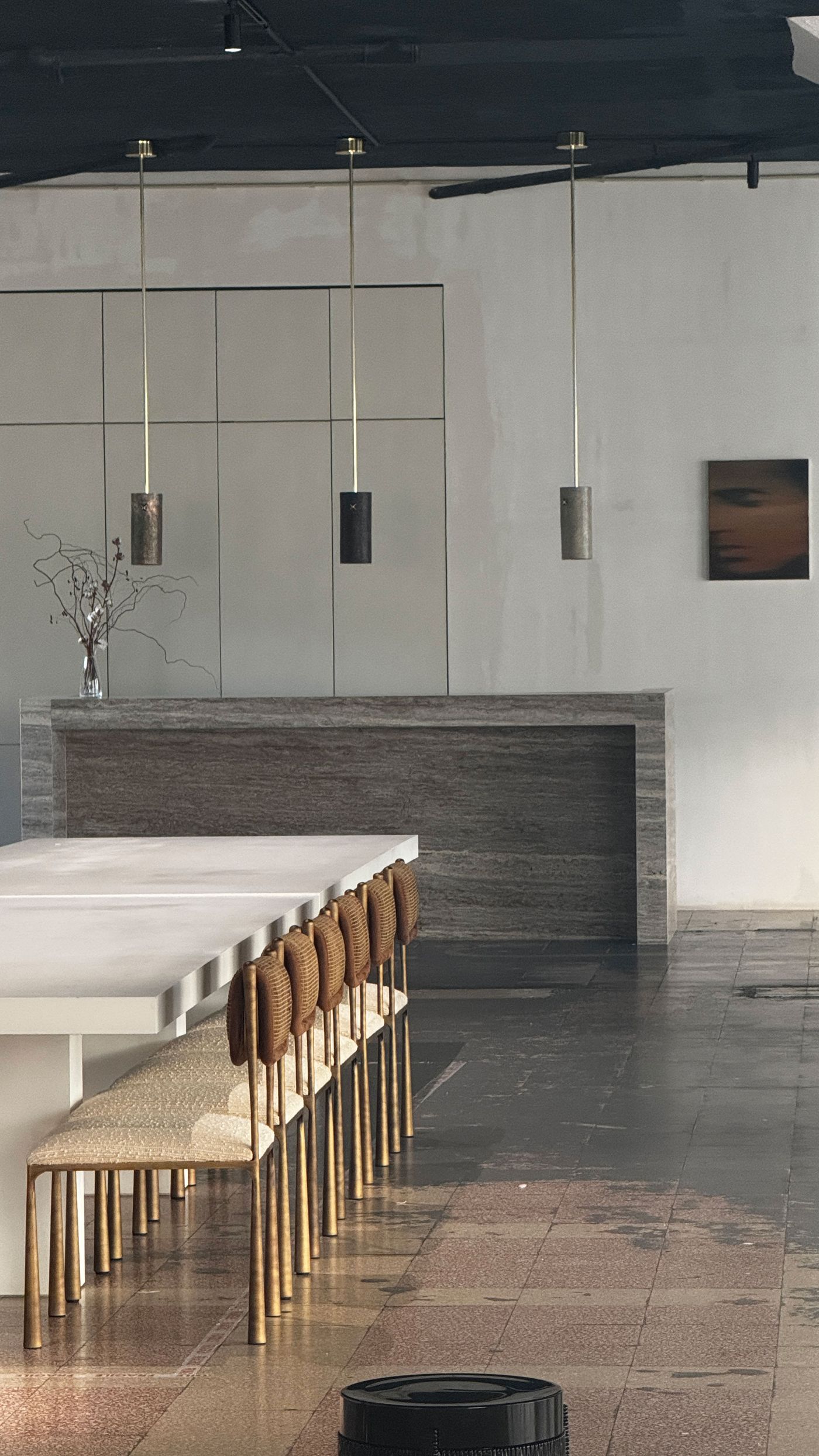 A contemporary dining or workspace by Hamrei features a long, low white table and chairs with slender gold legs and brown textured seatbacks. In the background, a substantial dark grey marble counter runs beneath sleek, minimalist cabinetry, with three slim cylindrical pendant lights hanging above. The space balances rich materials and clean lines, embodying a sophisticated, modern aesthetic.