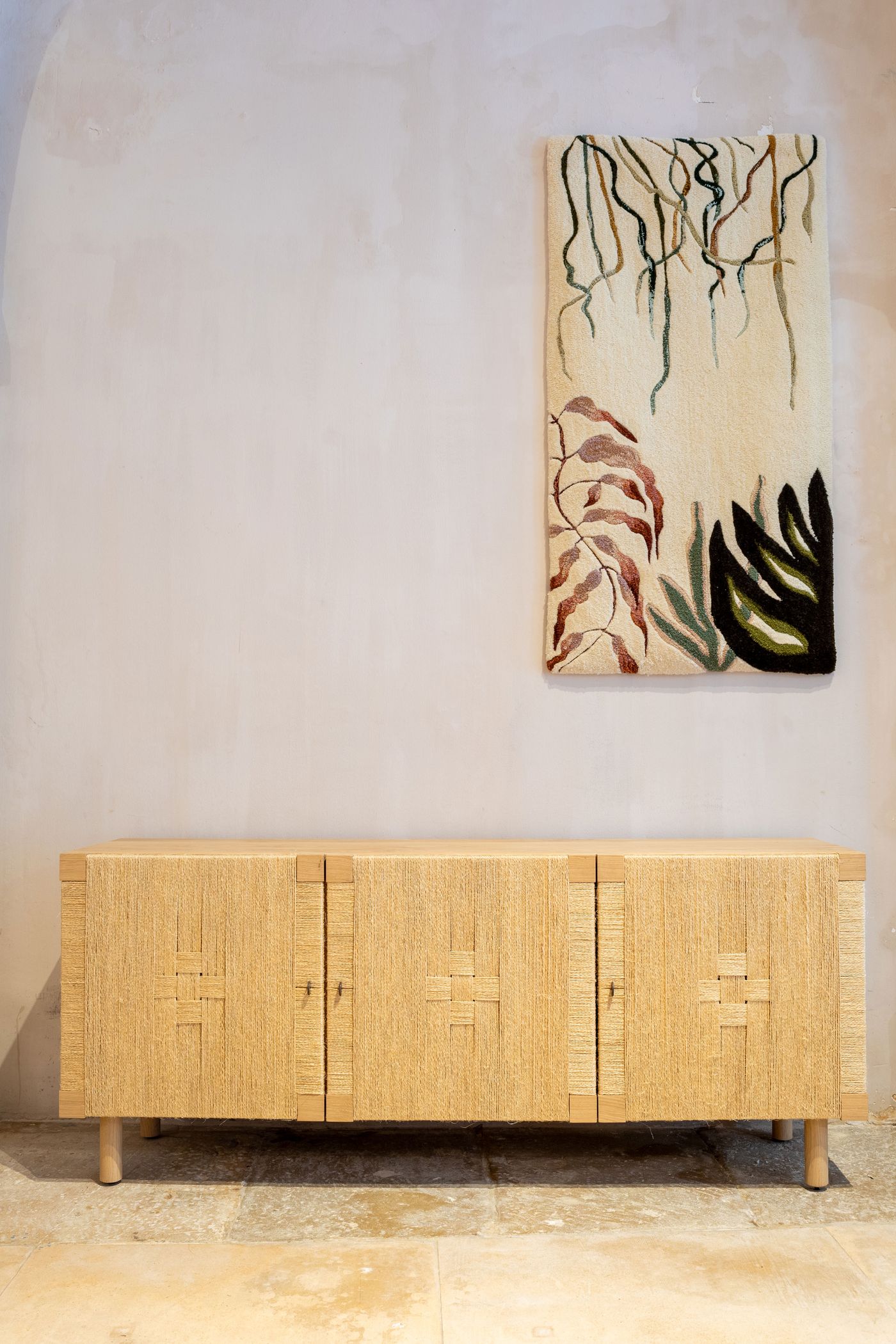 A natural-toned console cabinet, meticulously crafted with intricate woven straw patterns, stands below a vibrant abstract textile artwork depicting organic, flowing forms and leaves. The piece by Flores Textile Studio embodies biophilic and artisanal design, contrasting textural warmth with the minimalist wall, reflecting a thoughtful blend of craft and contemporary aesthetics.