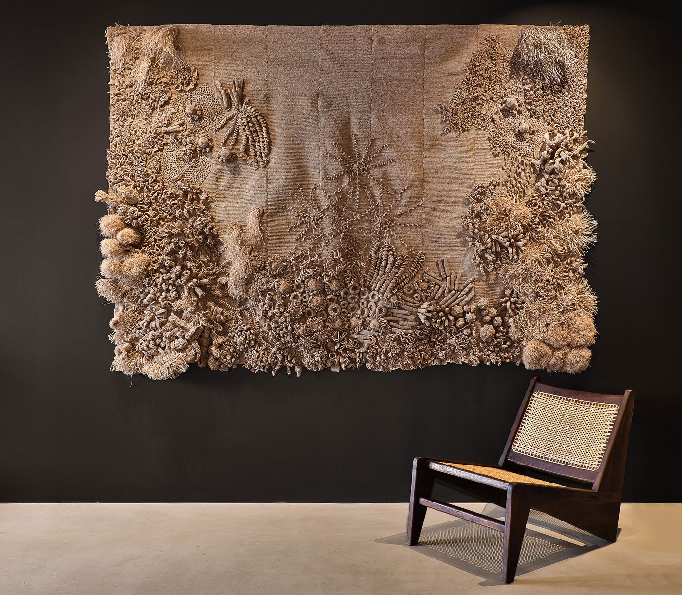 A captivating textured textile artwork, resembling organic forms and coral, dominates a dark wall in the Estadobruto showroom by Natalia Brilli Studio. Composed of natural fibers in varying shades of beige and brown, its intricate three-dimensional surface invites tactile exploration. A classic dark wood chair with a woven cane seat sits thoughtfully in the foreground, grounding the artistic expression.