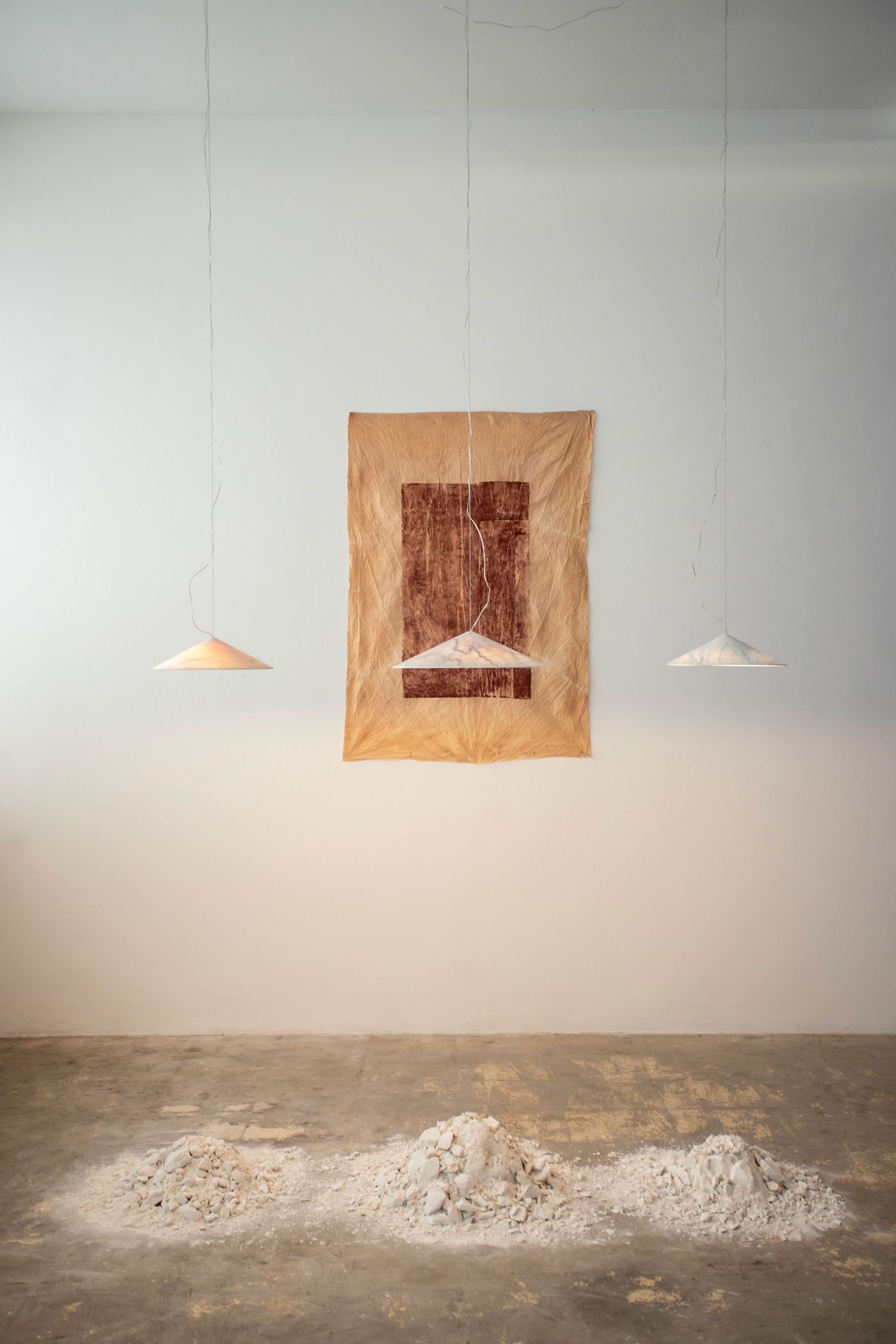 An enigmatic art installation features three suspended, conical pendant lights casting gentle glows. Behind the central light, a textured, earth-toned fabric tapestry with darker rectangular forms hangs against a white wall. Below, abstract piles of light-colored material rest on the concrete floor, creating a minimalist and contemplative scene that explores light, form, and raw materiality.