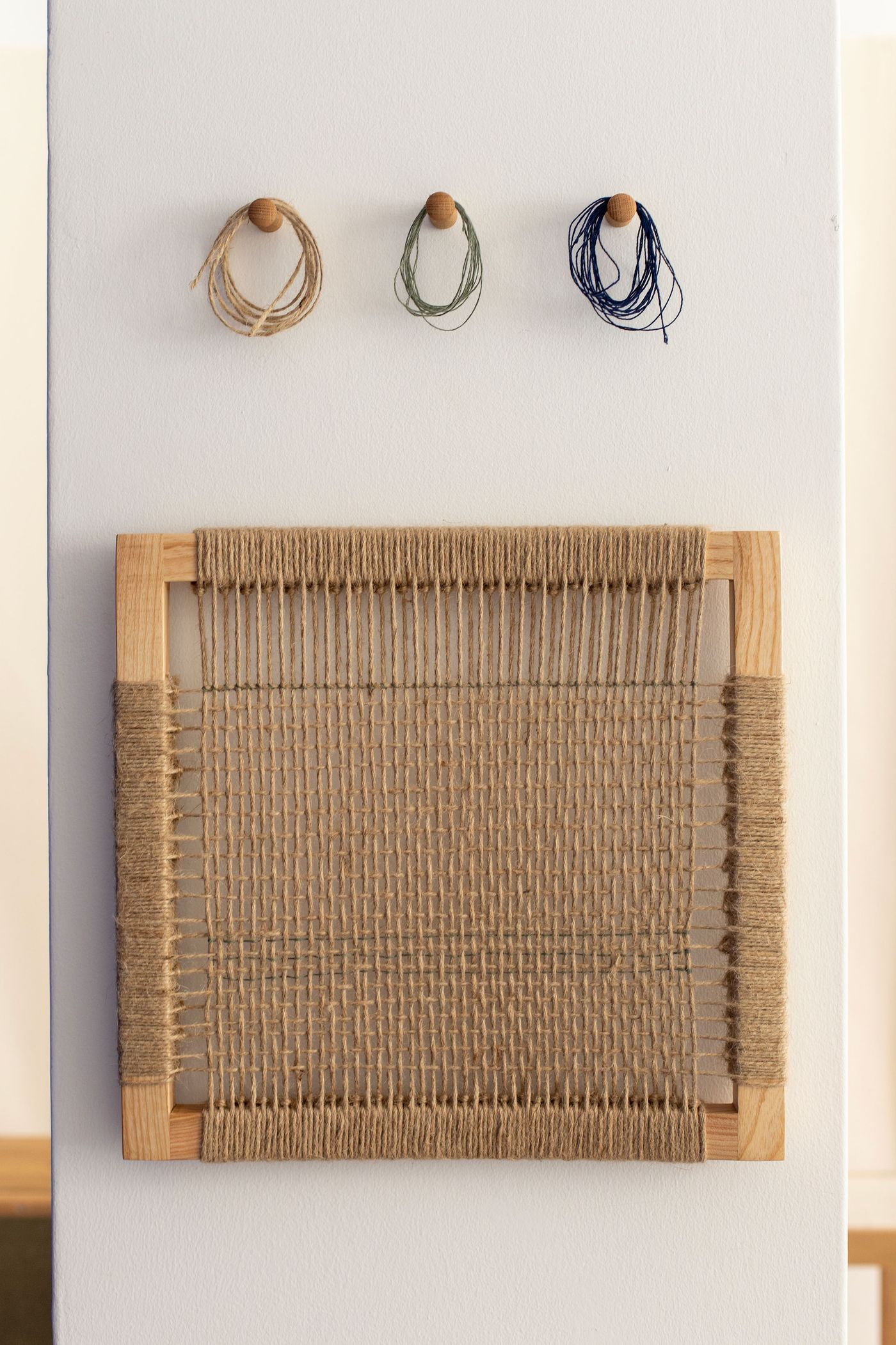 A minimalist wall display presents a square, raw wooden frame intricately woven with natural jute string, showcasing detailed textile craftsmanship. Above it, three small wooden pegs hold neatly coiled bundles of natural-toned, green, and dark blue twine, hinting at the creative process. The piece by Bamansure emphasizes organic materials and tactile artistry.