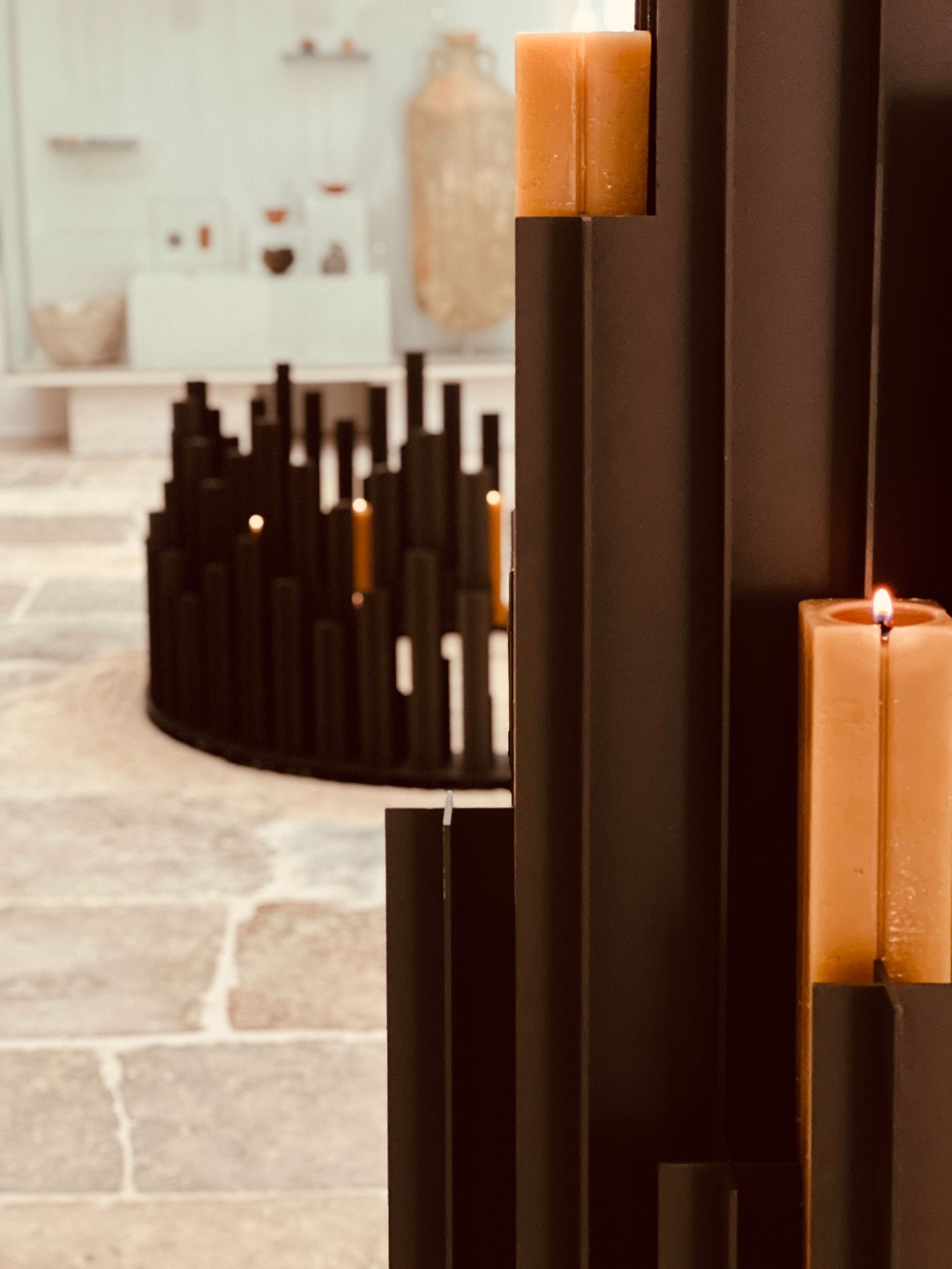 A captivating installation at Locke Santa Joana by AB and AC architects and Alma features two sculptural candle holders. In the foreground, dark, rectangular forms ascend, integrating amber block candles. In the mid-ground, a circular, fragmented dark form also holds lit candles. The rough stone floor contrasts with the modern structures, creating a mood of contemporary contemplation.