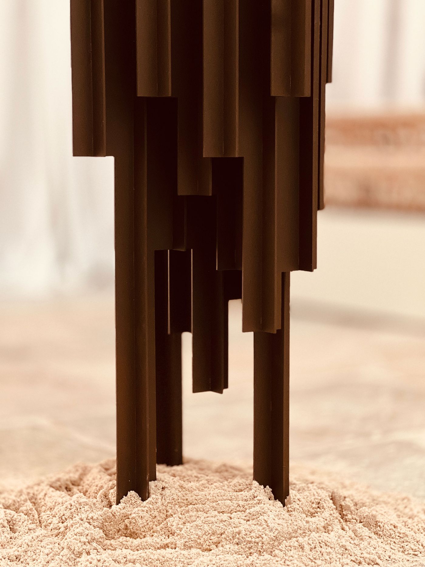 A close-up captures the base of a dark, sculptural object from AB and AC architects at Locke Santa Joana. Its stacked, interlocking rectangular forms descend into a bed of fine, pale sand or grit, suggesting a dialogue between structured design and natural elements. The image highlights textural contrast and the interplay of form and ground.