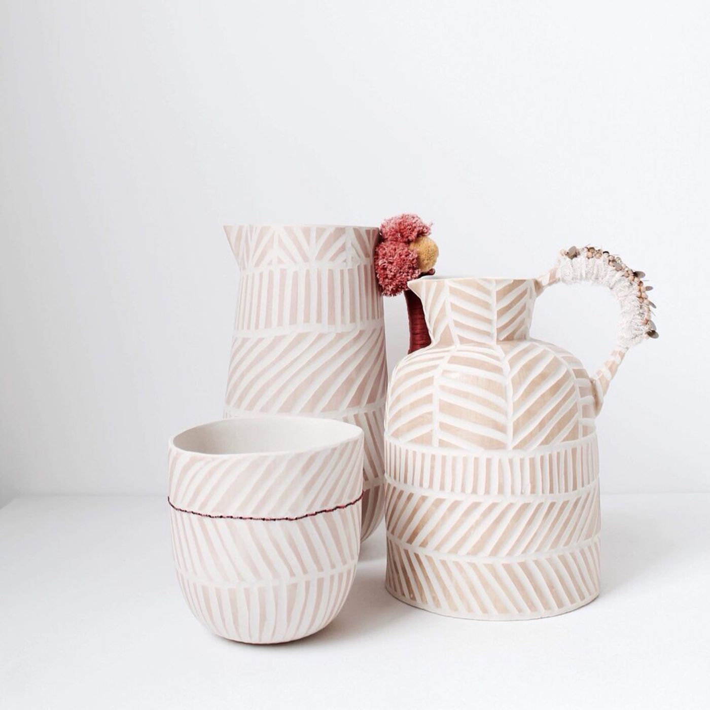 Three ceramic vessels by Anna Westerlund are arranged against a stark white background: a goblet, a pitcher, and a jug with an ornate handle. Each piece features a natural, light beige base adorned with a distinctive pattern of parallel, off-white lines, some angled, reflecting a modern take on traditional craftsmanship with subtle bohemian accents.
