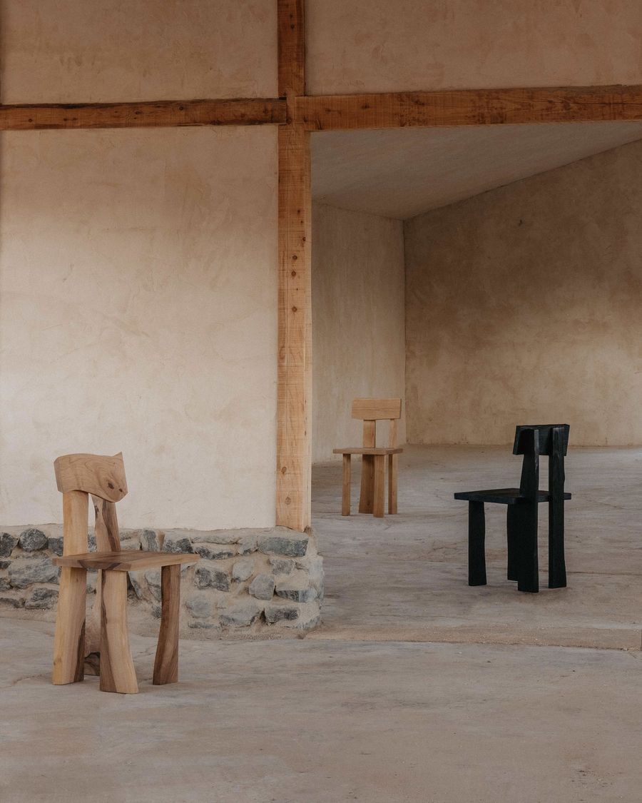 An expansive, raw architectural space with exposed wooden beams and a stone-based wall features three distinct, sculptural chairs by Ther. In the foreground, a light wooden chair with a carved back resembling an animal's head stands near a dark, blocky chair. The composition highlights unique, handcrafted forms in a rugged, neo-vernacular setting.