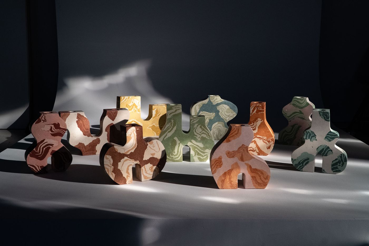 A collection of modern, sculptural vases from Tosco Studio's "Time Vessels" series, presented in various sizes and organic shapes. Each vase features a unique marbling effect in muted earth tones, soft greens, blues, and yellows, with some displaying cut-outs. Dynamic shadows cast across the white surface highlight their tactile forms and artistic craftsmanship.