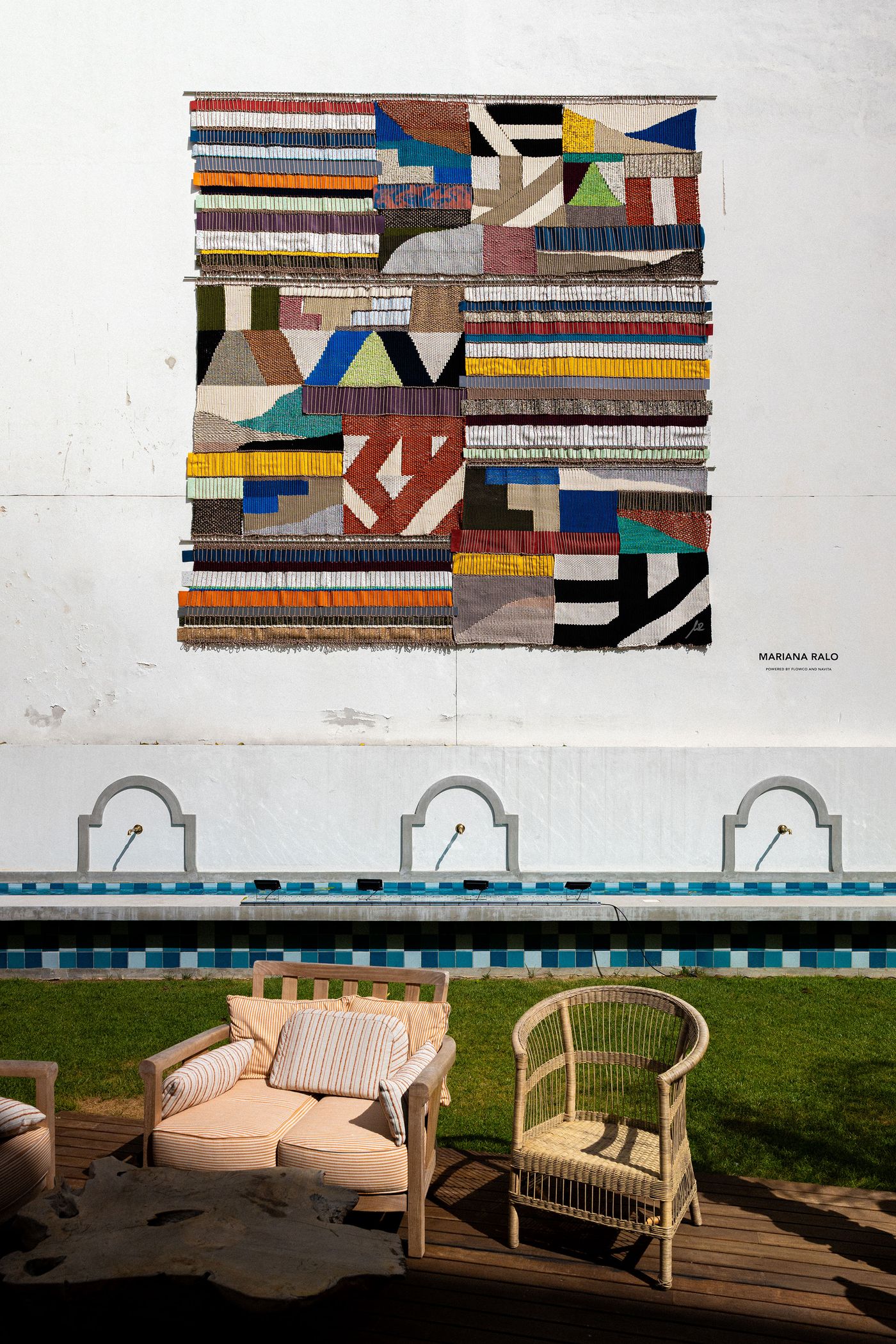 A vibrant textile artwork by Mariana Ralo hangs prominently on a textured white wall at Locke Santa Joana, showcasing a rich mosaic of woven colors and abstract patterns. Below, an outdoor seating area features two wooden chairs, one with an upholstered cushion and the other woven, positioned beside a long blue-tiled water feature with arching spouts, reflecting a blend of artistry and serene outdoor living.