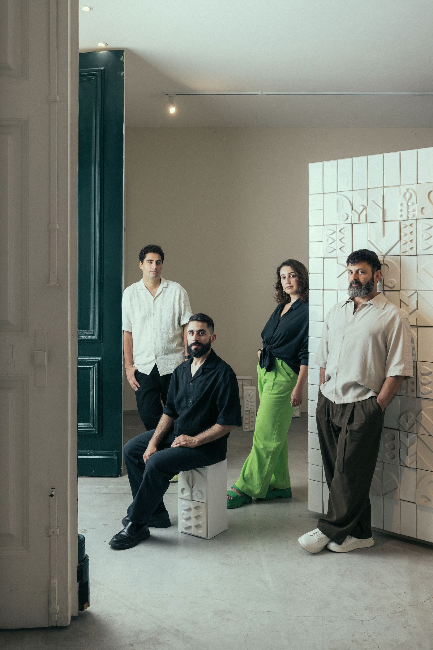 Four designers from Oficina Marques and Spacegram Studios are gathered in a bright, minimalist space, featuring a dark green doorframe and a white tiled architectural element with geometric relief. The group, dressed in modern, relaxed attire, exudes a collaborative and artistic energy, embodying the spirit of contemporary design and creative partnership.