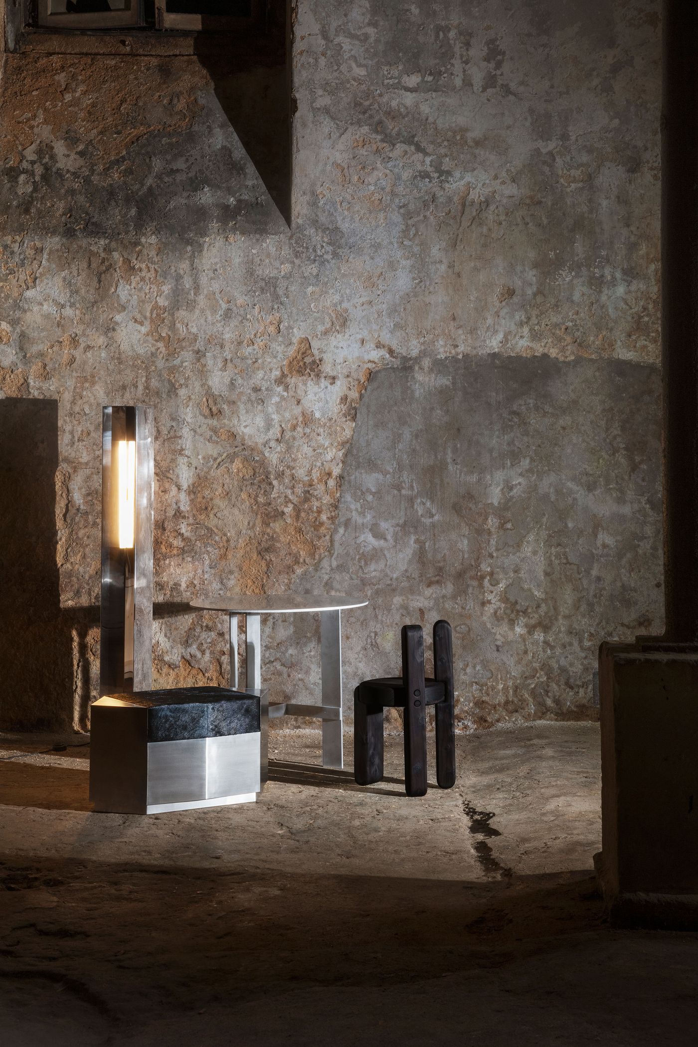In the raw, atmospheric setting of the Luso Collective's "Perspective Matter" exhibition, an illuminated modern floor lamp with a stark, linear form stands beside a minimalist white table and a dark, sculptural chair composed of block-like elements. The interplay of light and shadow on the textured concrete walls and floor enhances the dramatic mood, highlighting the contemporary pieces within a rustic, almost brutalist, architectural shell.