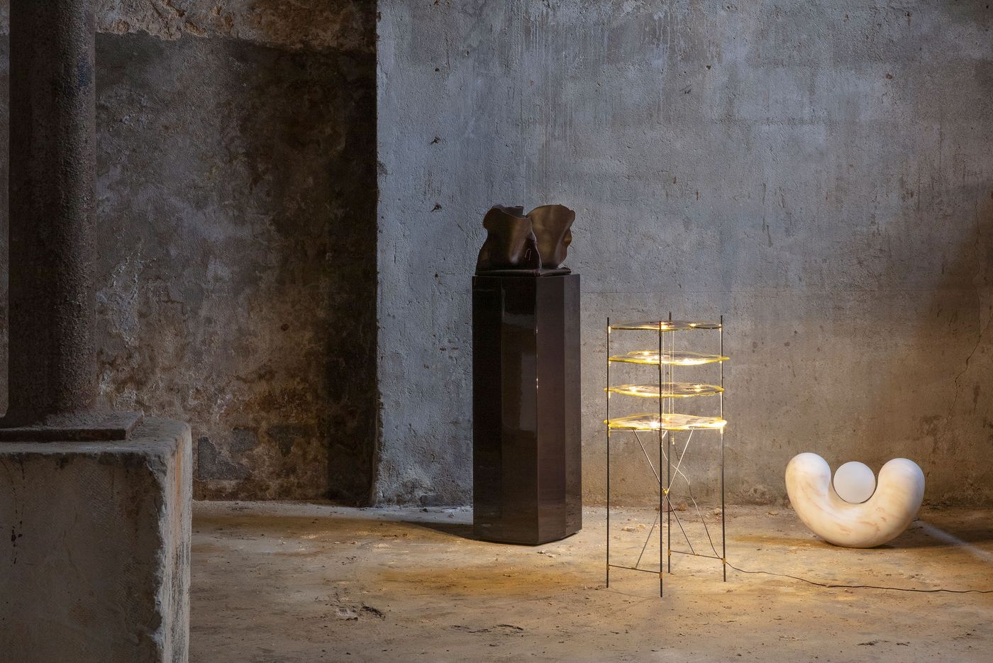 Within the raw, industrial setting of the Luso Collective's "Perspective Matter" exhibition, a dark, columnar pedestal displays a bronze, abstract bust. Nearby, a delicate, multi-tiered light sculpture with illuminated disc-like elements stands beside a smooth, organically shaped marble sculpture. The scene, bathed in soft, focused light, juxtaposes raw architecture with refined artistry, creating a contemplative atmosphere.