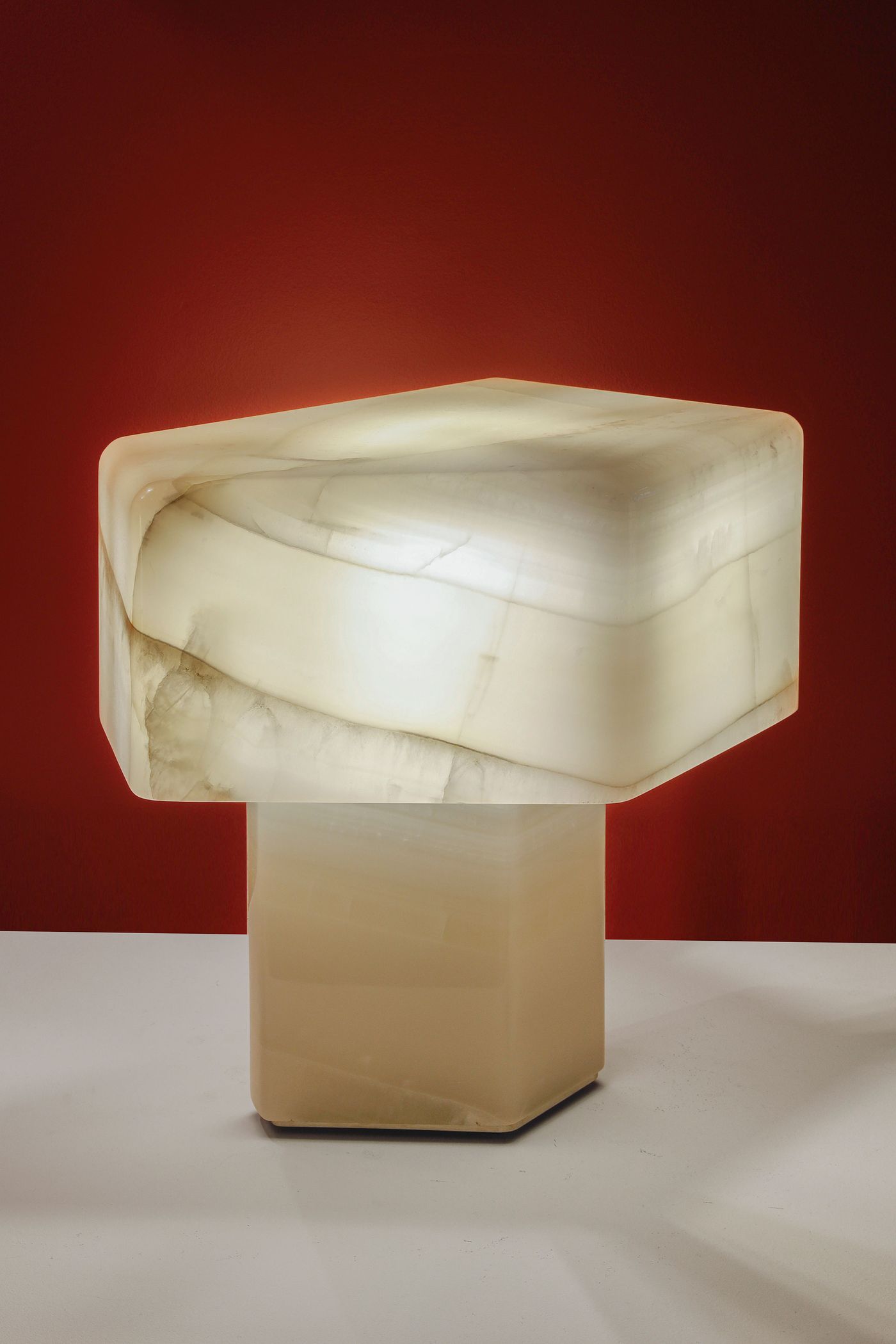 A mesmerizing light sculpture by Estadobruto, crafted from translucent, marbled white onyx, emanates a soft glow against a striking red backdrop. Its blocky, geometric upper form rests upon a more solid, rectangular base, showcasing the natural veining and luminous quality of the stone. This piece exemplifies a minimalist approach to lighting, transforming natural materials into ethereal art.