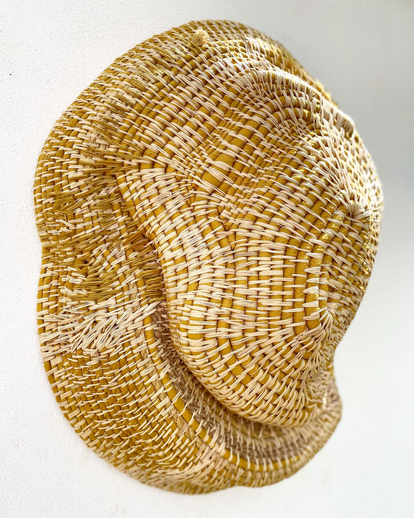 A sculptural wall art piece by Maria Pratas, intricately woven from natural fibers in a striking pattern of light cream and warm yellow. The organic, undulating forms create a sense of movement and depth, highlighting the artistry of traditional weaving techniques and the beauty of natural textures.