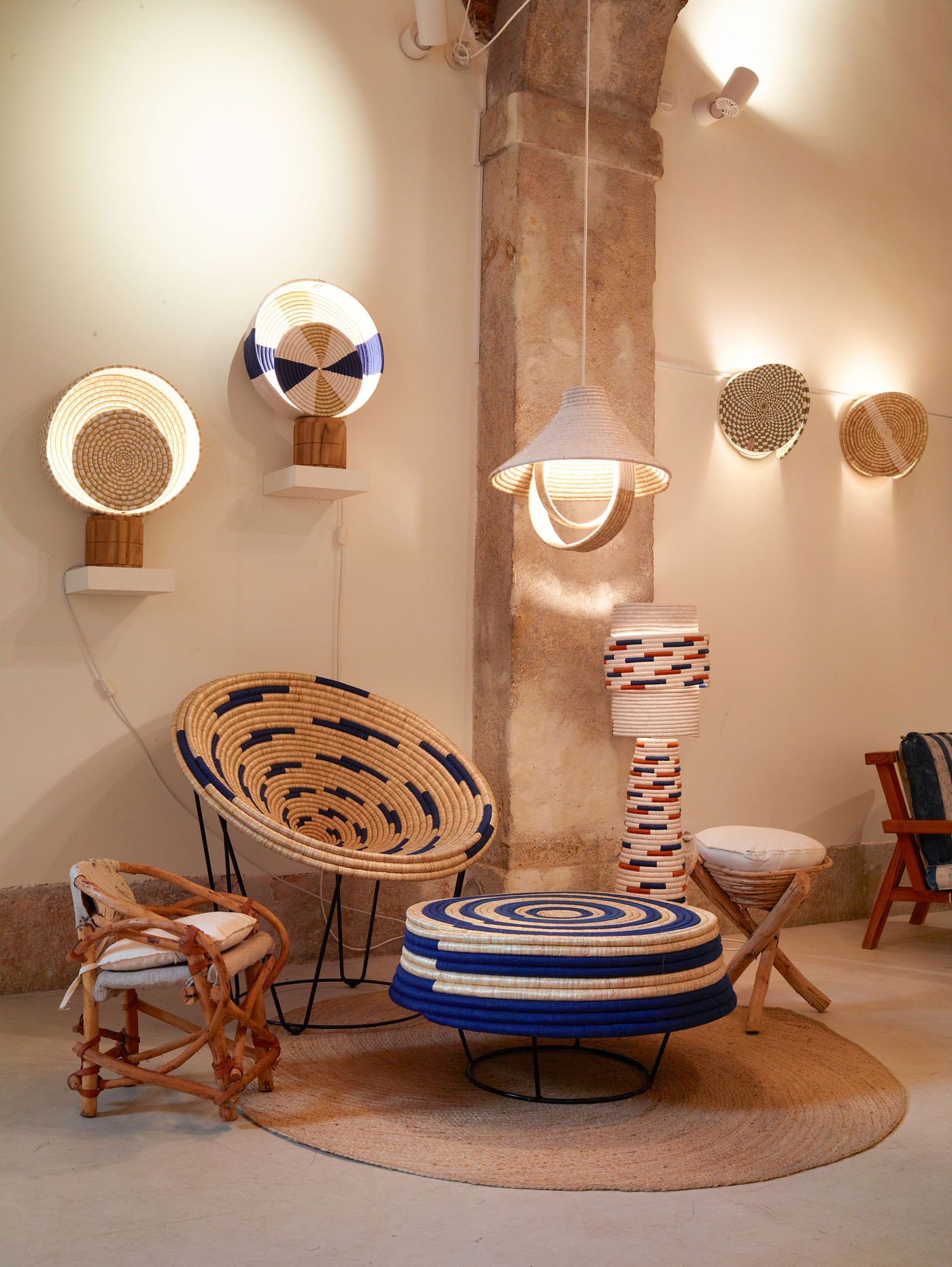 The Prime Matter Gallery's "Kukua" exhibition by Babled Studio features a rich display of woven natural fiber lighting and seating, set against a warm, neutral wall with an exposed architectural column. Circular wall-mounted lamps emit a warm glow, complementing a large round woven chair and a low table with concentric blue and natural patterns. The collection exudes a bohemian and artisanal aesthetic, emphasizing texture and handcrafted forms.