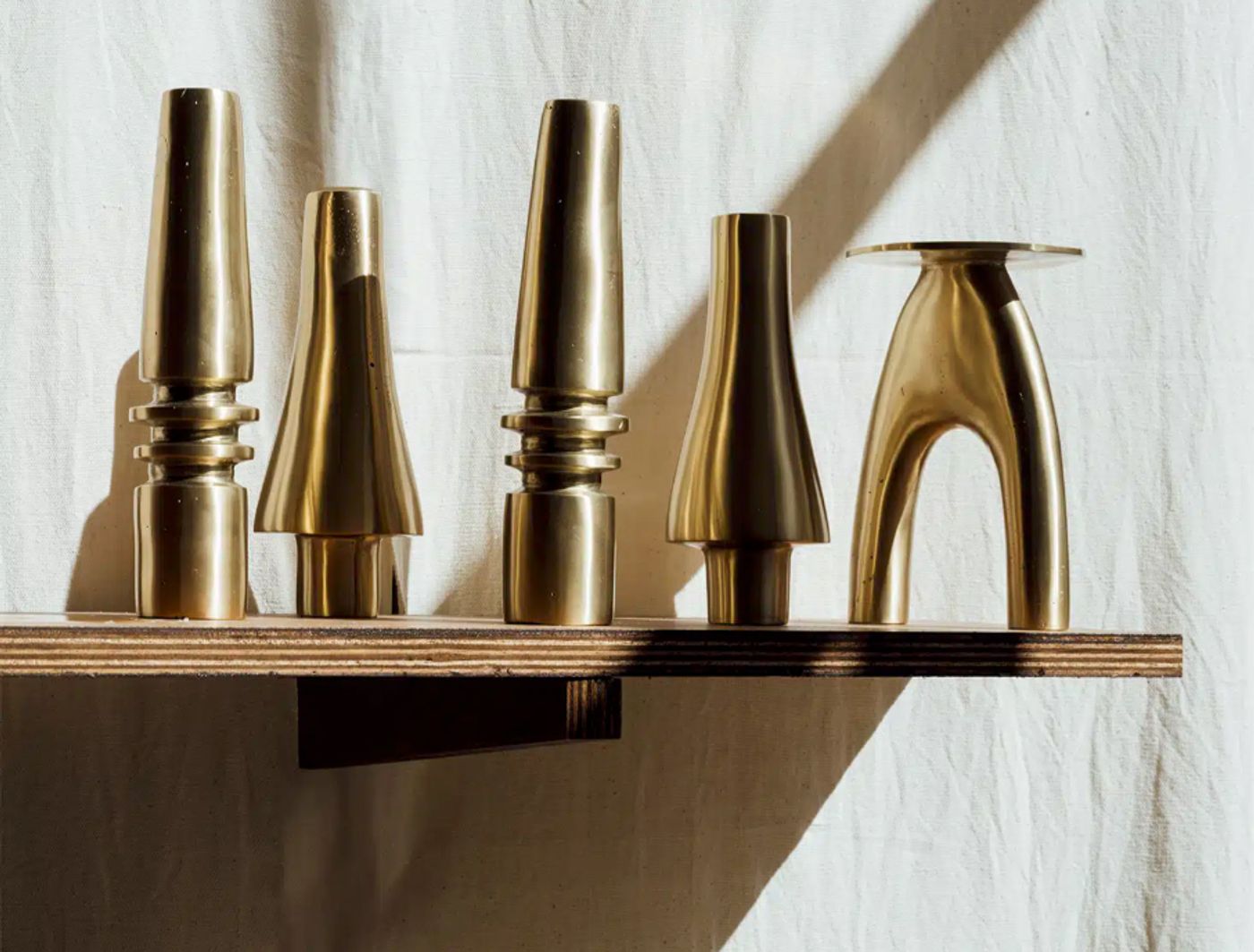 A collection of five abstract, highly polished brass sculptures from Maison Intègre are arranged on a light wooden shelf against a softly draped white fabric. Their forms range from conical to more architectural, with subtle rings and curves, showcasing a modern, refined aesthetic. The play of light and shadow accentuates the warm metallic sheen and the elegant simplicity of their shapes.