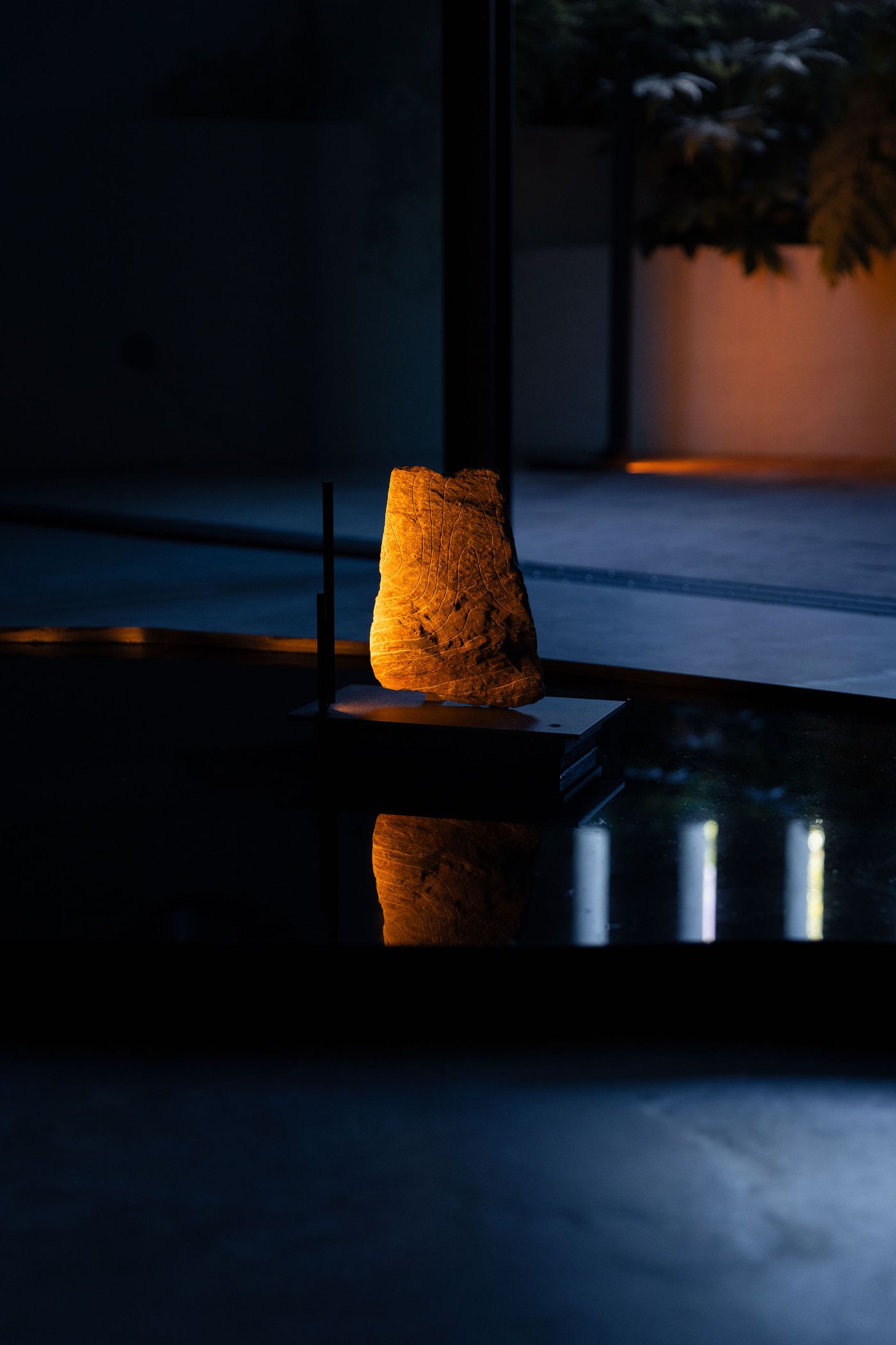 A softly illuminated raw stone sculpture by Made In Situ is prominently displayed on a dark, reflective surface, creating a striking vertical reflection. The warm light emanating from within the stone highlights its natural textures and organic form, creating a contemplative atmosphere. The ambient lighting and dramatic shadows emphasize the object's raw beauty as a focal point.