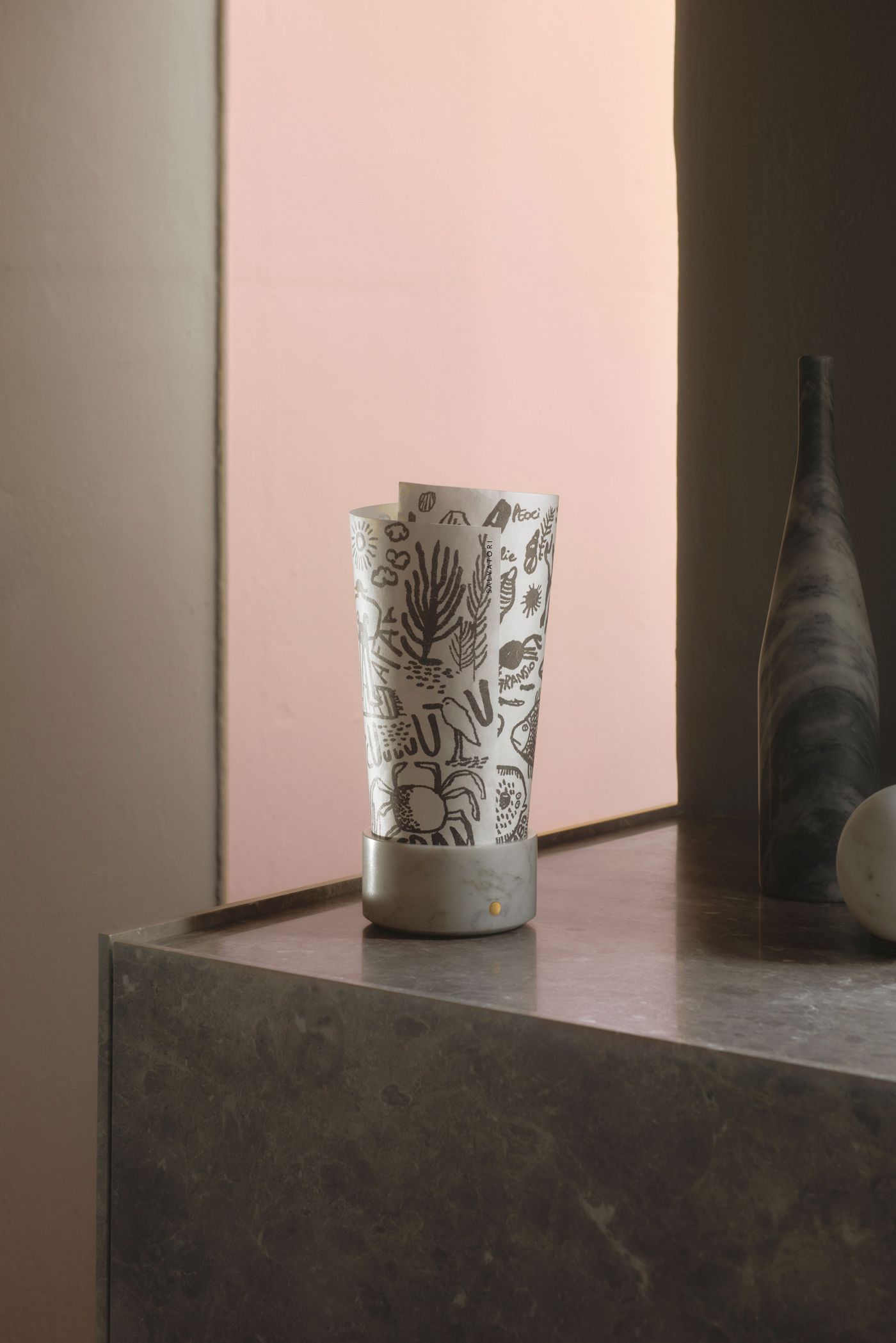 A sculptural lamp with a hand-drawn patterned paper shade and a marble base set against a pink wall.