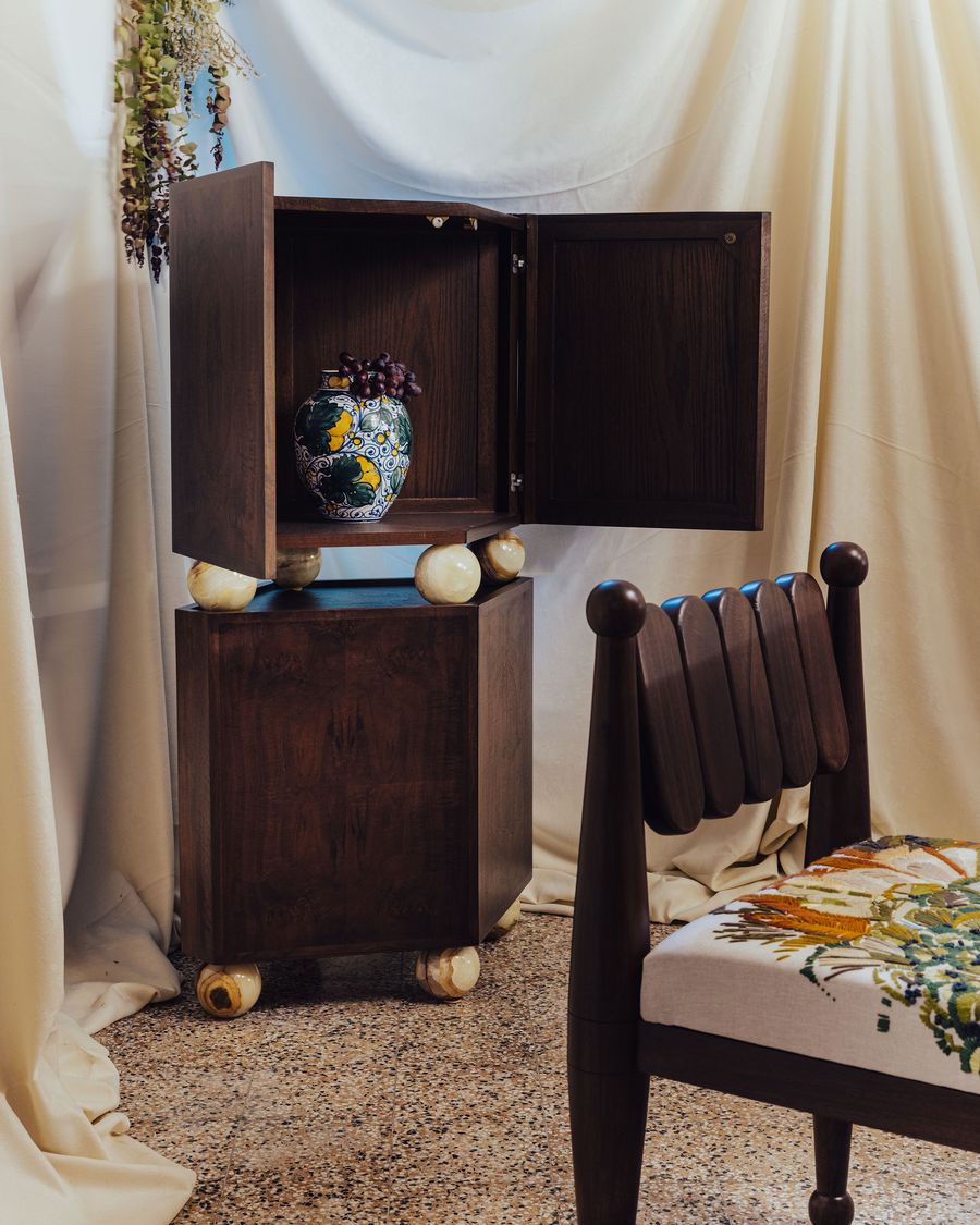 A sculptural wooden cabinet with marble-like spherical feet, paired with a dark wood chair upholstered in lush, nature-inspired embroidery, set against soft draped fabric — pure artisanal poetry. A sculptural wooden cabinet with marble-like spherical feet, paired with a dark wood chair upholstered in lush, nature-inspired embroidery, set against soft draped fabric — pure artisanal poetry.