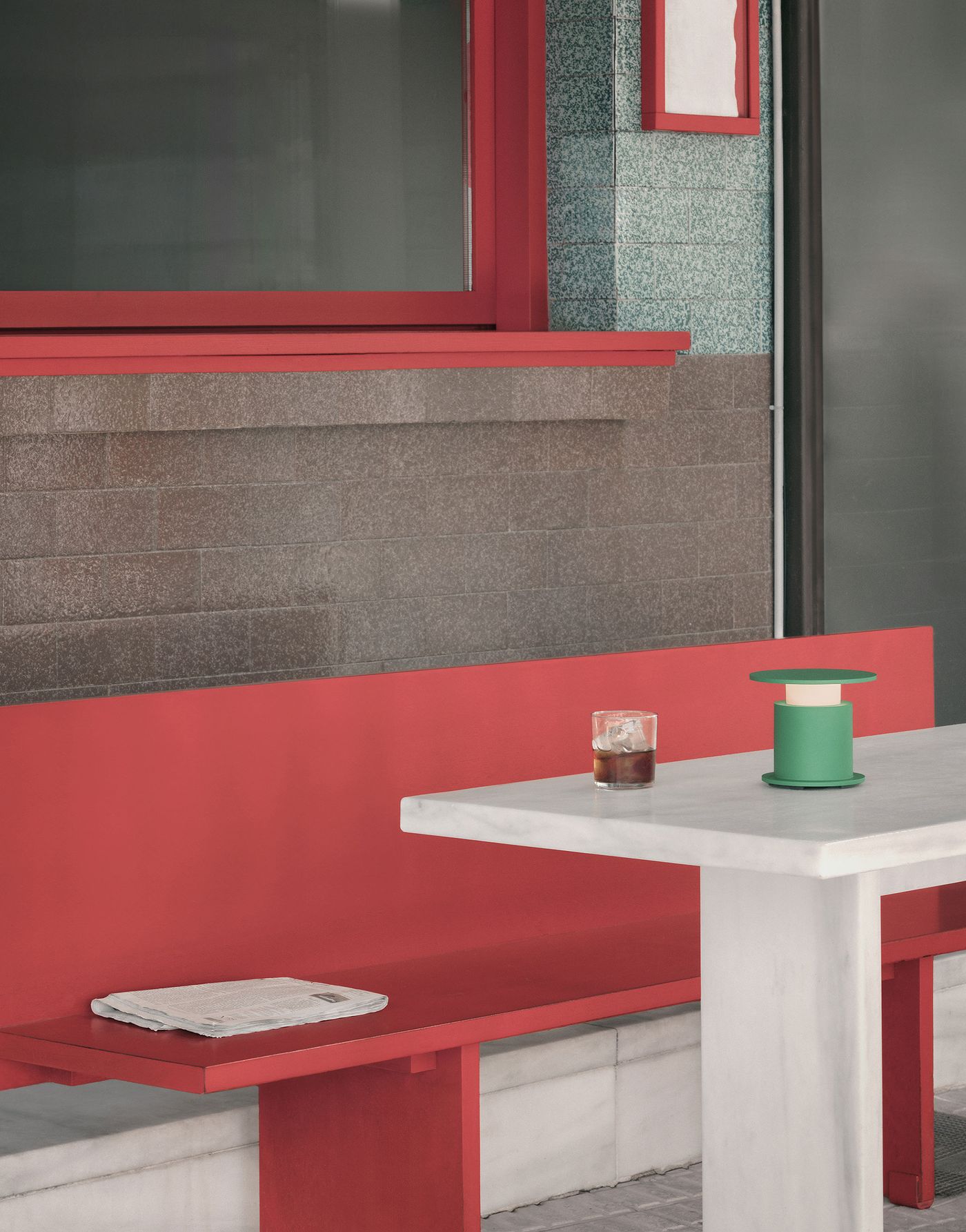 A vivid green version of the w241 Faro table lamp brightens a marble-topped table in a vibrant outdoor café with red benches and a folded newspaper.