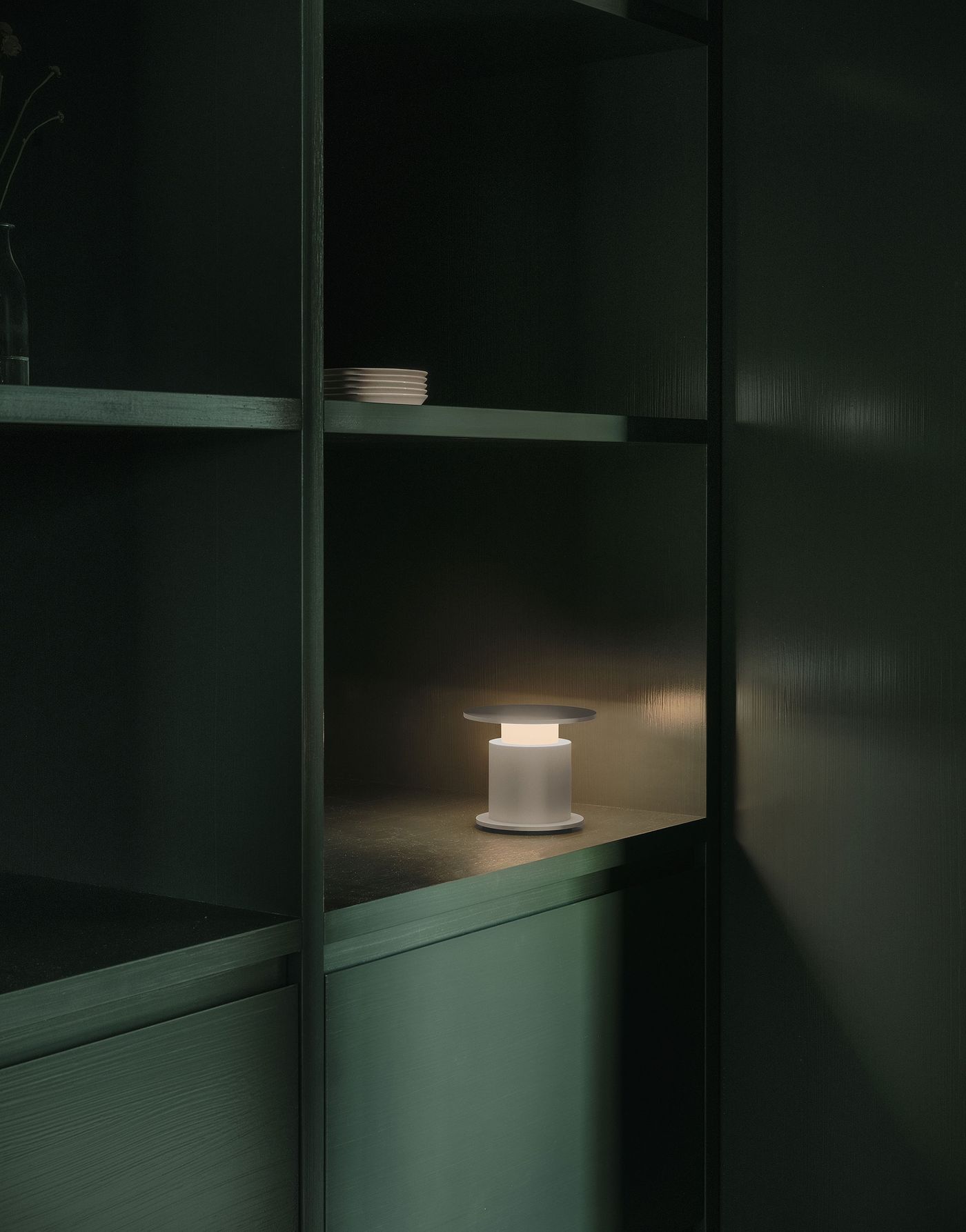 A soft-lit w241 Faro table lamp with a flat disc top placed on a dark green built-in shelf, casting a warm glow in a minimalist interior.